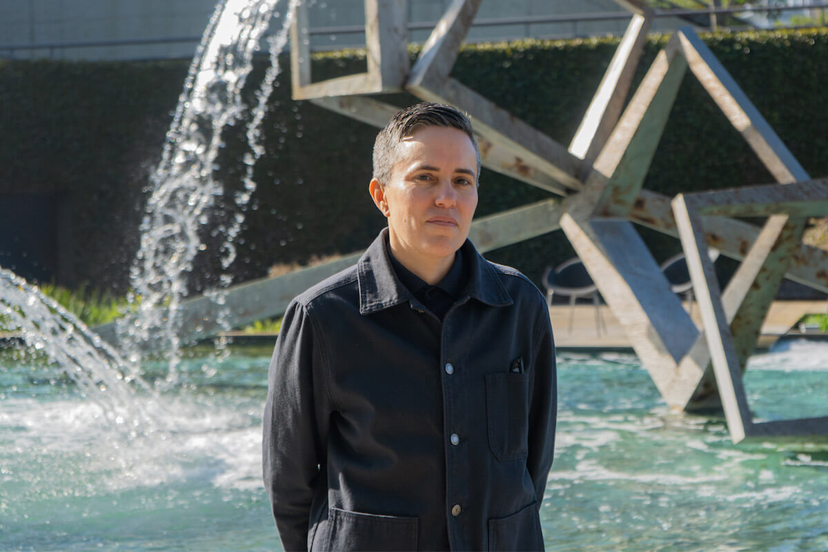Person in a dark jacket standing beside a cascading fountain and angular metal sculpture.